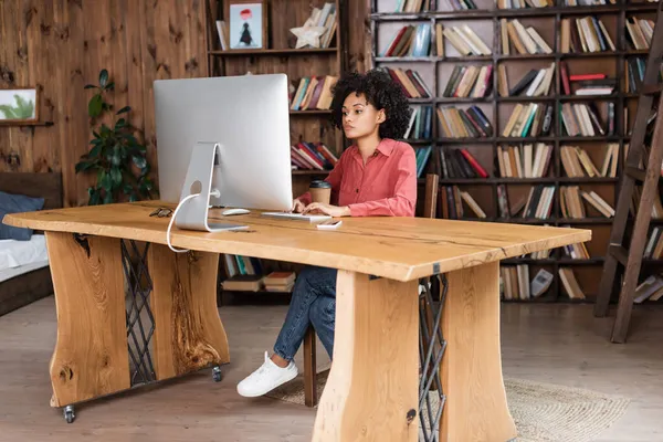 Jovem afro-americana digitando no teclado do computador enquanto olha para o monitor em casa — Fotografia de Stock