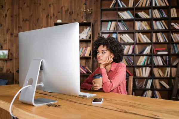 Кучерява афроамериканська жінка дивиться на монітор комп'ютера і тримає паперову чашку вдома — стокове фото