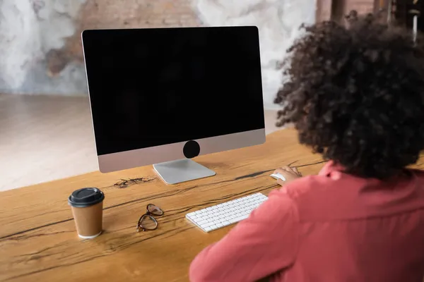 Back view of curly african american woman using computer near paper cup and eyeglasses on desk — Stock Photo