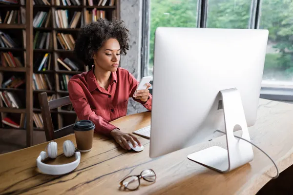 Mujer afroamericana rizada usando teléfono inteligente cerca del monitor de la computadora, taza de papel y auriculares en el escritorio - foto de stock