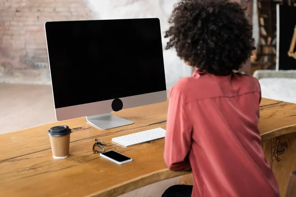 Vue arrière de femme afro-américaine bouclée utilisant un ordinateur près du smartphone, tasse en papier et lunettes sur le bureau — Photo de stock