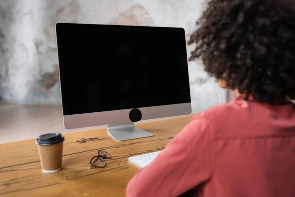 Back view of blurred african american woman near computer monitor, paper cup and eyeglasses on desk — Stock Photo