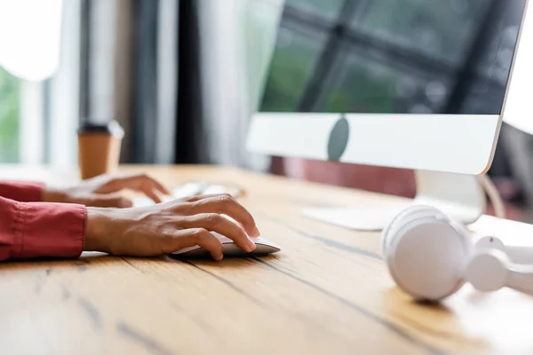 Vue recadrée de femme afro-américaine à l'aide d'une souris d'ordinateur près d'une tasse en papier floue et d'écouteurs sur le bureau — Photo de stock