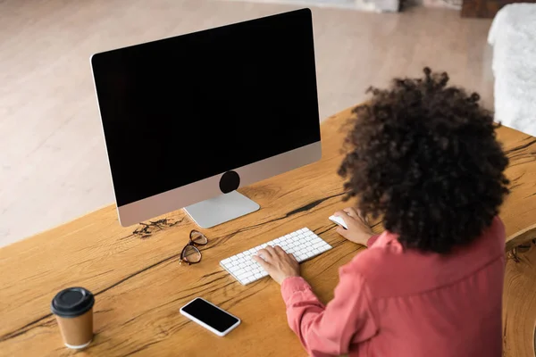 Vista de ángulo alto de la mujer rizada afroamericana utilizando el teclado de la computadora cerca de teléfono inteligente, taza de papel y anteojos en el escritorio - foto de stock