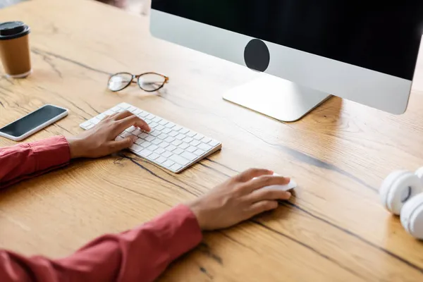 Vista recortada de la mujer afroamericana utilizando el teclado de la computadora cerca de teléfono inteligente, taza de papel y gafas en el escritorio - foto de stock