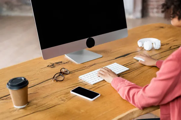 Vue partielle d'une femme afro-américaine utilisant un clavier d'ordinateur près d'un téléphone portable, une tasse en papier et des lunettes sur le bureau — Photo de stock