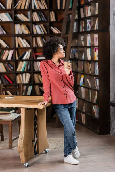 Mujer afroamericana rizada sosteniendo taza de papel cerca del escritorio - foto de stock