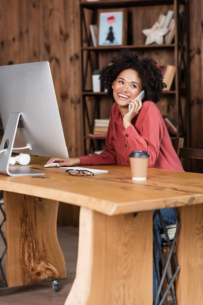 Mulher americana africana alegre falando no smartphone perto do monitor, copo de papel e óculos na mesa — Fotografia de Stock