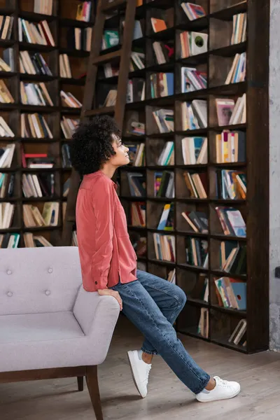 Vista lateral de la mujer afroamericana apoyada en un sofá gris cerca de la estantería - foto de stock