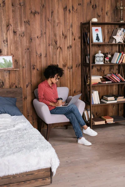 Jovem freelancer afro-americano sentado em poltrona com laptop e segurando copo de papel — Fotografia de Stock