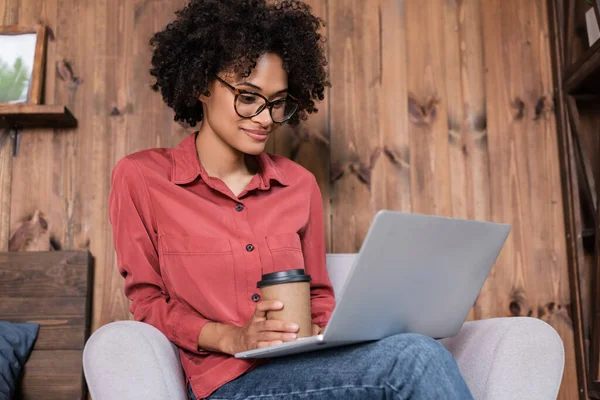 Felice freelance americano africano utilizzando il computer portatile e tenendo tazza di carta mentre seduto sulla poltrona — Foto stock