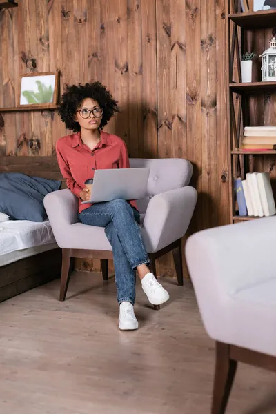 Freelance africano americano pensoso usando il computer portatile e tenendo tazza di carta mentre seduto sulla poltrona — Foto stock