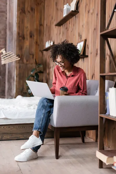 Freelancer afro-americano alegre usando laptop e segurando copo de papel enquanto sentado em poltrona — Fotografia de Stock