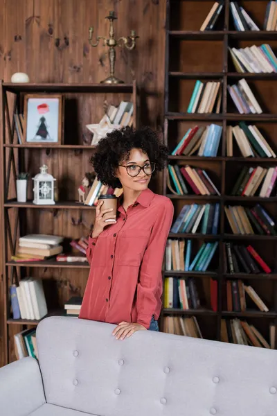Cheerful african american woman holding paper cup while standing near couch — Stock Photo