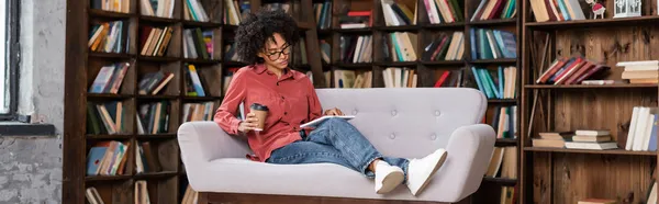 Lockige afrikanisch-amerikanische Frau hält Pappbecher in der Hand, während sie ein digitales Tablet in der Nähe des Bücherregals benutzt, Banner — Stockfoto