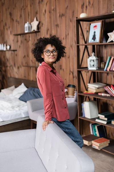 Happy african american woman holding paper cup while standing near couch — Stock Photo