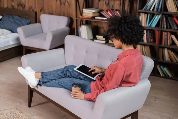 Mujer afroamericana rizada sosteniendo una taza de papel mientras usa una tableta digital - foto de stock