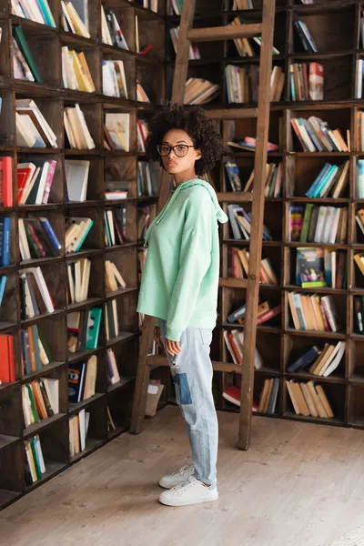 Junge afrikanisch-amerikanische Studentin mit Brille steht neben Bücherregal und Leiter — Stockfoto