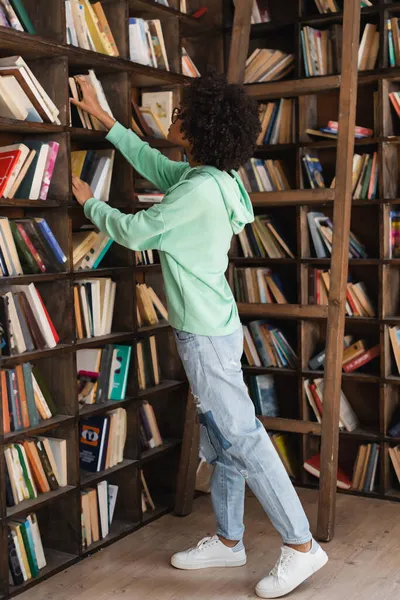 Jovem estudante afro-americano em óculos atingindo livros em estante perto de escada — Fotografia de Stock