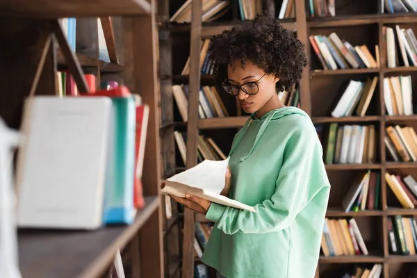 Молодий афроамериканець у окулярах читає книжку біля розмитої книжкової шафи на передньому плані. — стокове фото