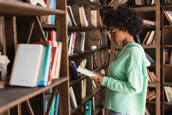 Vista lateral do estudante americano africano em óculos livro de leitura perto estante borrada em primeiro plano — Fotografia de Stock
