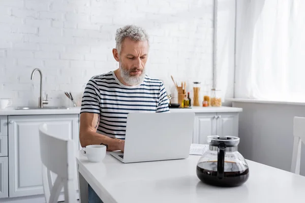 Homme mûr en t-shirt rayé utilisant un ordinateur portable près du café dans la cuisine — Photo de stock