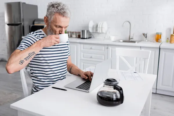 Man drinking coffee and using laptop near papers in kitchen. Translation: 