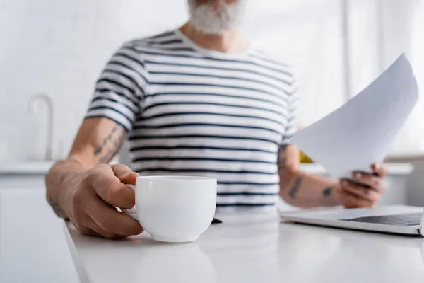 Обрізаний вид чашки кави в руці розмитого чоловіка з паперами біля ноутбука вдома — стокове фото