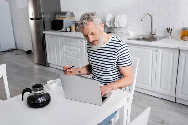 Hombre maduro sosteniendo pluma y el uso de ordenador portátil cerca de café y olla en la cocina - foto de stock