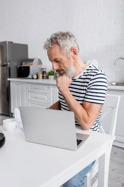 Homem maduro barbudo olhando para o papel perto de laptop e café na cozinha — Fotografia de Stock