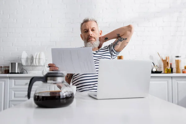 Hombre maduro sosteniendo papeles cerca de la computadora portátil y cafetera borrosa en la cocina - foto de stock