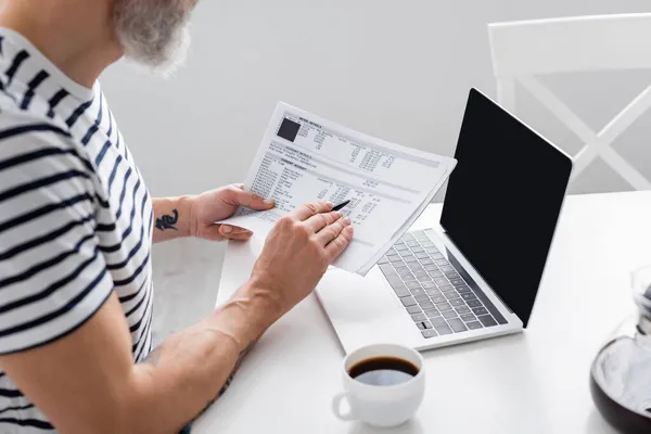 Cropped view of bearded man holding bills near laptop with blank screen and coffee in kitchen — Stock Photo