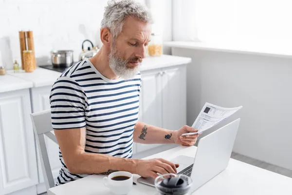 Homem maduro segurando papéis com contas e usando laptop perto de xícara de café na cozinha — Fotografia de Stock