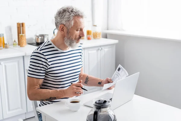 Vista lateral do homem de meia idade segurando papel com contas e caneta perto de laptop e café em casa — Fotografia de Stock