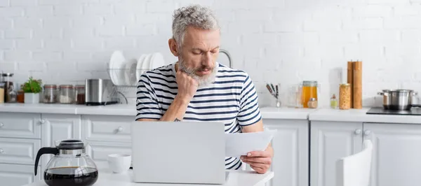 Зрелый мужчина в полосатой футболке с бумагой рядом с ноутбуком и кофейником на кухне, баннер — стоковое фото