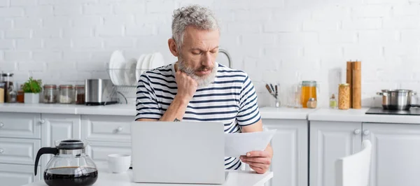 Mature man in striped t-shirt holding paper near laptop and coffee pot in kitchen, banner — Stock Photo