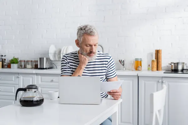 Homem de meia idade olhando para o documento perto de café e laptop na cozinha — Fotografia de Stock