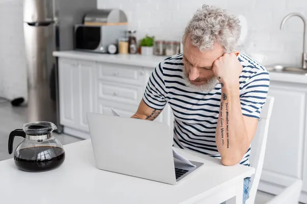 Trauriger Mann mit Dokumenten neben Laptop und Kaffee in der Küche. Übersetzung: 