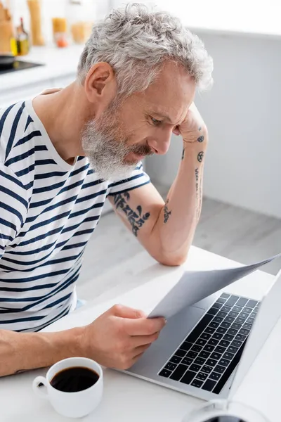 Hombre maduro tatuado sosteniendo papeles cerca de la computadora portátil y café en casa - foto de stock