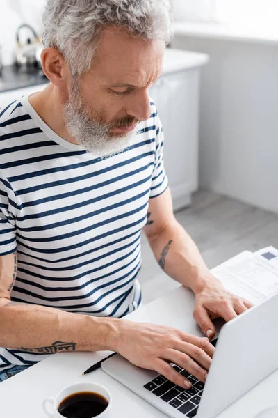 Man using laptop near coffee and papers in kitchen. Translation: 