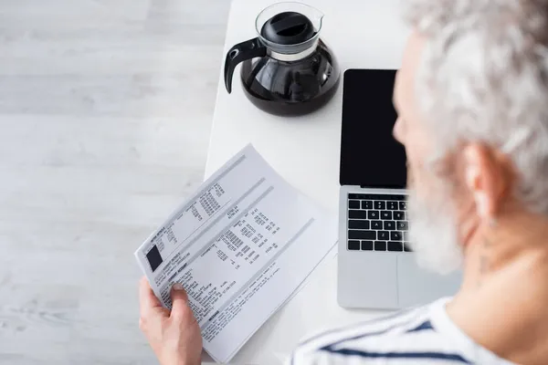 Blurred mature man holding papers with bills near laptop and coffee pot — Stock Photo