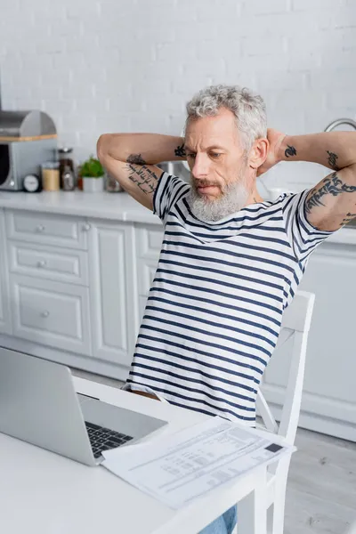 Uomo tatuato guardando laptop vicino a carte con fatture in cucina — Foto stock