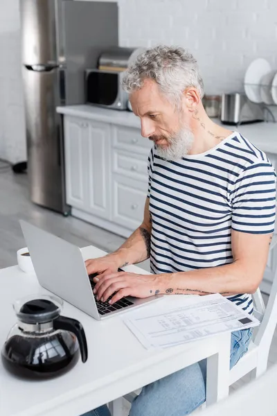 Mature man using laptop near documents with bills and coffee at home — Stock Photo