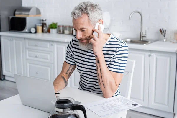 Hombre hablando en el teléfono inteligente y el uso de ordenador portátil cerca de facturas y café en casa. Traducción: 