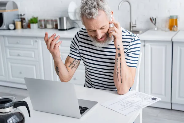 Lächelnder älterer Mann spricht auf Smartphone und schaut in der Küche auf Scheine in der Nähe des Laptops — Stockfoto