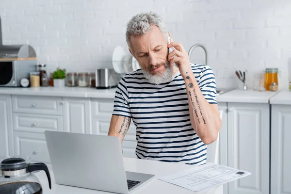 Tattooed mature man talking on mobile phone near bills and laptop in kitchen — Stock Photo