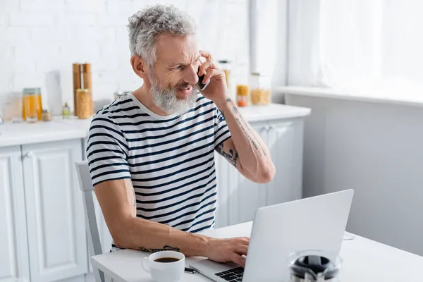 Freelancer maduro positivo falando no smartphone e usando laptop perto de café na cozinha — Fotografia de Stock