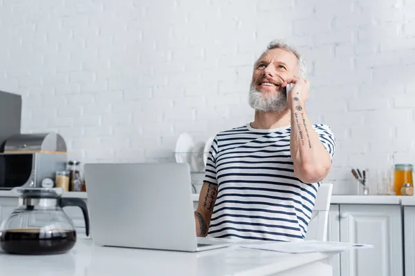 Freelancer positivo hablando en el teléfono móvil cerca de la computadora portátil y cafetera en la cocina - foto de stock