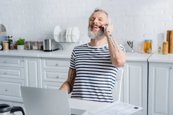 Happy mature man talking on smartphone near laptop near bills in kitchen — Stock Photo