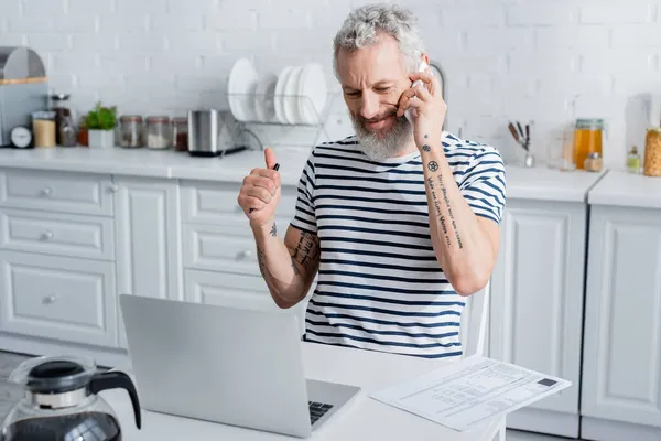 Hombre tatuado mostrando como mientras habla en el teléfono inteligente cerca de facturas y portátil en la cocina - foto de stock